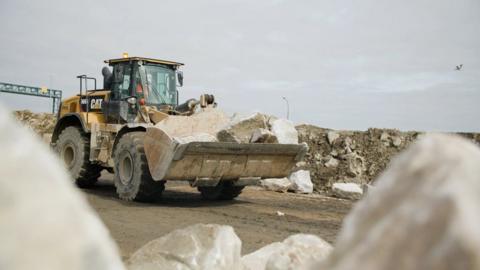 A yellow digger carries rocks along a road surrounded by other large rocks.