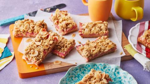 A chopping board with an assortment of oat crumble bars sitting on top of it, next to some cups and bowls.