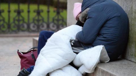 A homeless person is sat on a step wearing a thick winter coat and carrying a sleeping bag.