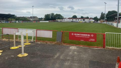 The pitch at Frome Town surrounded by stands, with a red gate in foreground.