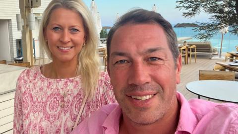 Anna Voyce stands behind Tom Voyce in a holiday location with the sea behind. She is wearing a pink dress and has blonde hair. He has darker hair and is wearing a pink shirt.