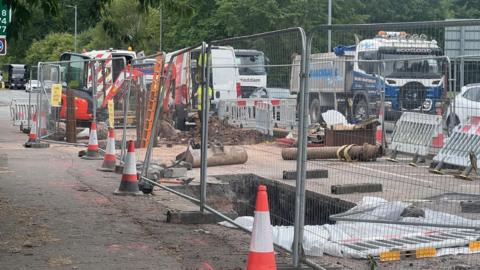 A fenced off hole in the ground with cones around it and traffic passing by behind it