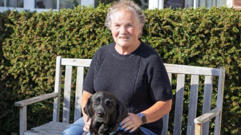 A woman sitting on a bench with a black guide dog beside her.