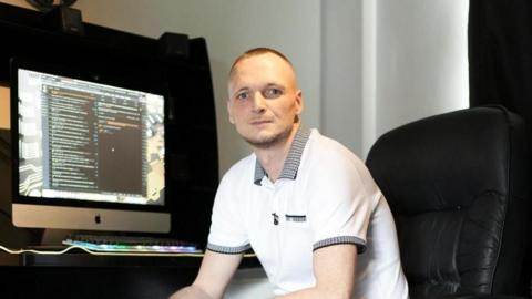 A man with short hair, wearing a white polo shirt, is sat in a large black office chair and in front of a computer screen.