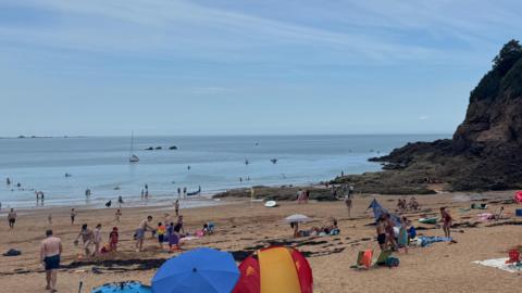 Busy sandy beach with tents and umbrellas, people relaxing and swimming, and a rocky cliff by the sea under a cloudy sky.
