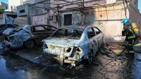 An image of a burnt-out blue car in front of a heavily damaged building with blown-out windows and burned exterior walls. A firefighter in uniform sprays the blue car