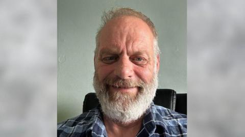 Simon Burke, a middle aged man with a white beard and hair. Wearing a blue checked shirt. 