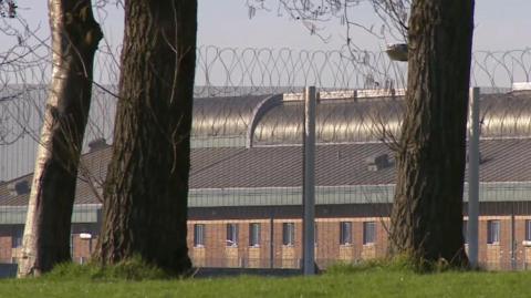 The prison surrounded by a security high fence and barbed wire and two trees in the foreground