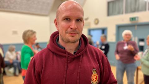 A bald-headed man with a red hoodie staring at the camera with women in the background chatting