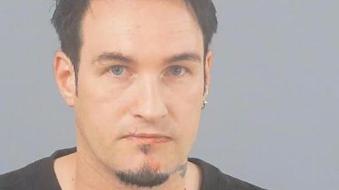 In a police custody photo, Ben Lewis has black hair, a black goatee beard, blue eyes and a small circular earring in one ear.