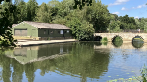 A river is pictured on a sunny day. The water is camp with a slight ripple going over it. Two people walk over a stone bridge in the background. A green building, which is a boat club, is seen across the water.