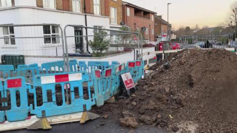 Temporary works in a road see a pile of mud and a hole in the ground, surrounded by temporary fencing.