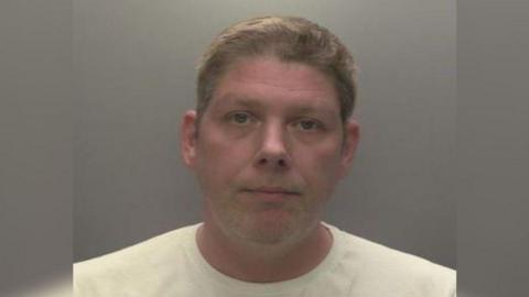 A police mugshot of Stephen Verrill, who is wearing a white t shirt and has  short brown hair. He is looking into the camera with a glum expression.