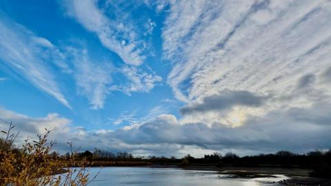 sun reflecting onto a lake, trees around the edge, fluffy and streaky cloud amidst some bright blue sky