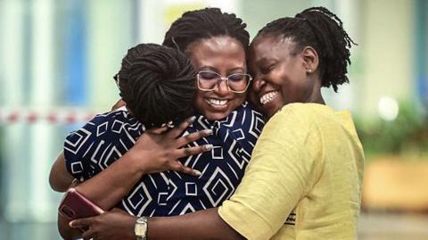 A person wearing a patterned dress is embracing two other people in an airport arrivals area, standing close together near a piece of luggage with a bag placed on top of it. The background shows a brightly lit terminal with glass walls and blurred interior details.