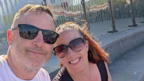 Craig and Trish Hinde smiling together for an outdoor selfie wearing sunglasses.