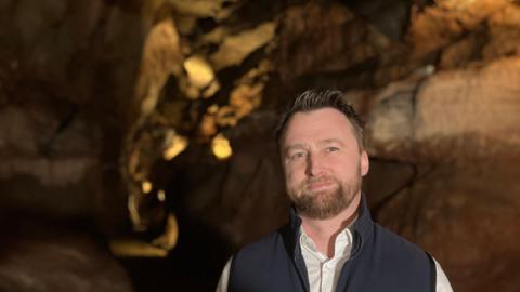 A man in a white shirt and blue fleece stands inside a cave, the stone walls are brown and lit up. 