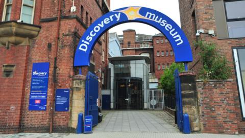 The entrance to the Discovery Museum. The venue's name is carried on an arched blue sign above opened metal gates. The building is largely built from brick but has a glass foyer.