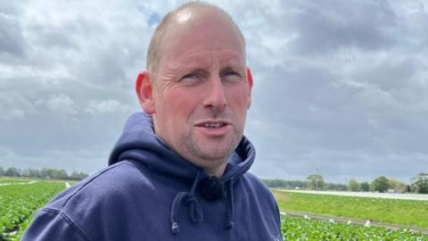 Jon, who is wearing a navy hoodie, is standing in a field of crops with his hands on his waist. 