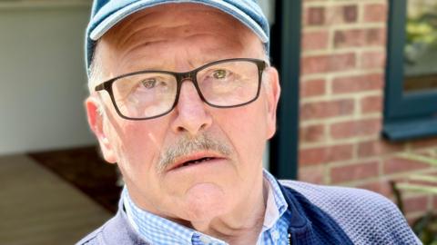 Close up photo of Ray sitting in the garden. He's wearing a blue baseball cap, black glasses and a blue shirt. He's looking into the camera with little expression on his face.