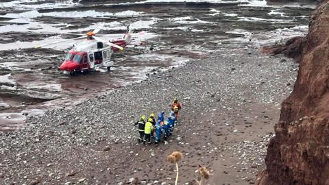 A groups of emergency services personnel - firefighters and coastguard crew - carrying a stretcher (which you cannot see). There is a red and white helicopter on the sand nearby