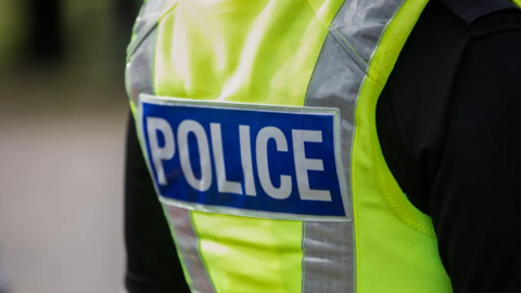 The back of a police officer who has a black top on with a yellow hi-vis vest on. It has silver reflective strips and it says "police" on it.