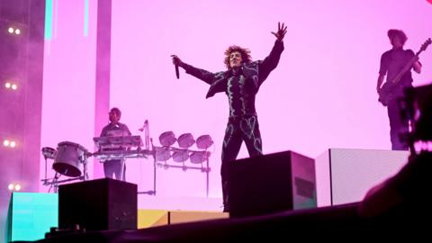 Three members of a rock band perform on a pink-lit stage. The lead singer stands with arms aloft and is holding a portable microphone. Behind him to the left of the picture a band member stands behind a keyboard and drums set and to the right is a man holding an electric guitar.