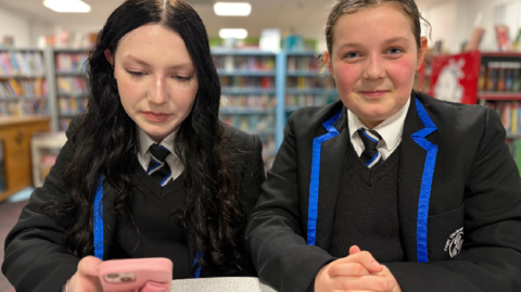 Two girls wearing school uniform with black blazers and blue edging standing together in a library with one holding a pink mobile