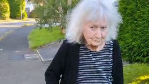 Anne Stacey walking down a pavement. The picture is from the waist up. She has grey bobbed hair and is wearing a striped navy blue and white top and a black cardigan. Driveways, lawns and trees can be seen in the background 