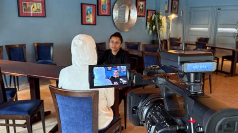 A person with their hood up speaking to a person across the table, with a camera in front of them filming the conversation. 