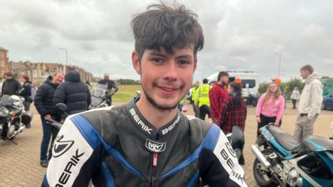 William wearing black and blue motorcycle leathers with short curly hair, smiling at the camera. There are people loitering in the background in a car park with a few other motorbikes in view.
