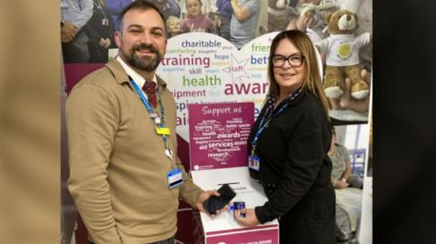 Ben wears a mustard colour jumper and has a beard, and Dawn is smartly dressed and wears glasses. The donation point is a cardboard post box, and they are holding their phone and credit cards next to it.