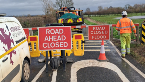 Road closed signs, traffic cones and a metal gate block a road with a flooded field next to it. A man in hi vis walks next to it and a white van is parked to the left.