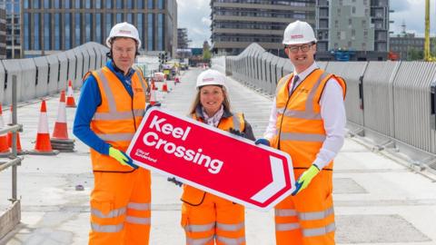 Councillor Kevin Johnston, councillor Lindsey Leonard and councillor Michael Mordey, hold a red sign reading Keel Crossing at the new footbridge. They are wearing orange protective clothing and white helmets. The grey concrete structure is still under construction. There are more new building on its other side, including the City Hall. Keel Square is also visible in the distance.