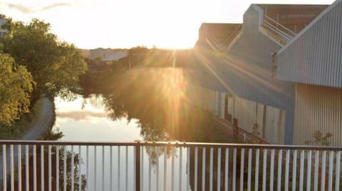 A canal is pictured at sunset from a bridge above. The camera looks over black and white railings. The river is about 30ft wide and looks calm. Trees grow on the bank to the left and are reflected in the water. Industrial warehouses stand on the right bank.