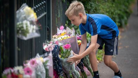 A boy wearing a blue shirt and black shorts is laying flowers on the ground next to other bouquets. They are by a school fence.