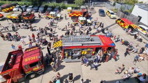 Aerial shot of a fire service open day, with people milling around an assortment of fire engines in a large concreted area.
