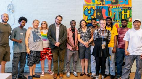 Twelve people stand in a line and are smiling at the camera. Fifth from the left is Matt Storey who is wearing square-shaped glasses, a grey blazer and brown trousers. He has short brown hair and dark facial hair. There is a colourful mural on the blue wall behind the group which has letters on it spelling: 'Youth.'