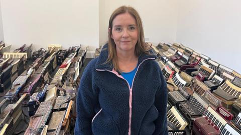 Lisa Allen standing in a room surrounded by dozens of accordions.  She has shoulder-length fair hair and is wearing a blue top under a navy fleece with a pink zip.  Accordions of many different designs are laid out on the floor in neat rows. 