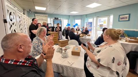 A group of people in the Shores Centre they are sat at tables having afternoon tea and clapping their hands