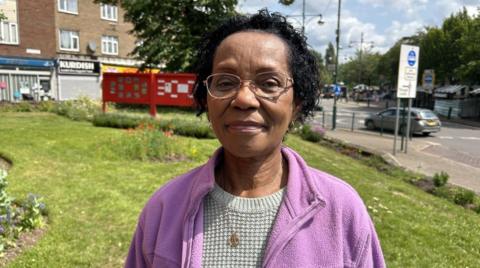 Norma Thompson in Wednesfield town centre. There are some shops and a car behind her. She has black hair and is wearing a jumper and purple fleece. She has a necklace on and is wearing glasses.