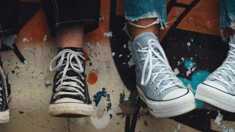 Two young people sit on a graffiti-wall with their Converse-style trainers hanging over it. The shot is focused on two pairs of trainers that are black and light blue. 