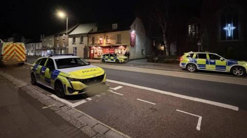 The picture shows a quiet street at night with a strong police presence. There are three marked police vehicles visible: a car parked in a designated bay in the foreground, another car across the street near a pub, and an SUV further down the road. A large police van is also parked on the left side of the image.