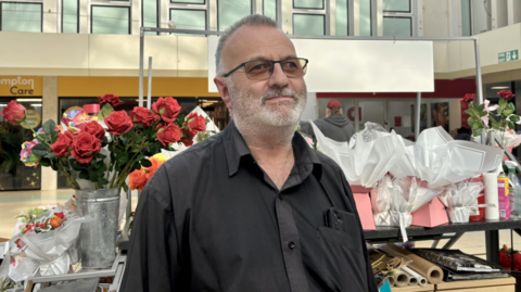 A man with a black shirt and glasses on standing in front of a flower stand. He has grey short hair and a bear and has a phone in his shirt pocket. 