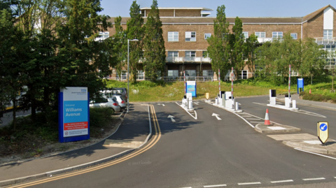 A screenshot from Google street view of the road entrance to a hospital. There's a barrier system on the road, a blue and white sign on the left hand side and a brick building in the background. It's a sunny day and the sky is blue