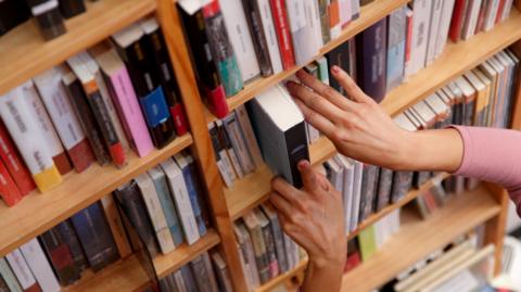 A woman's hands can be seen removing a book from a shelf of books in a library.
