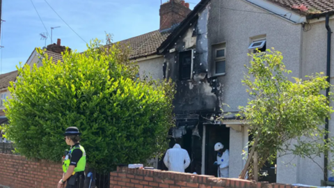 A fire-damaged house on a residential street. A police officer is standing in front of the property and two forensic officers are standing in the entrance. The entrance to the house and an upstairs window are burnt out. 