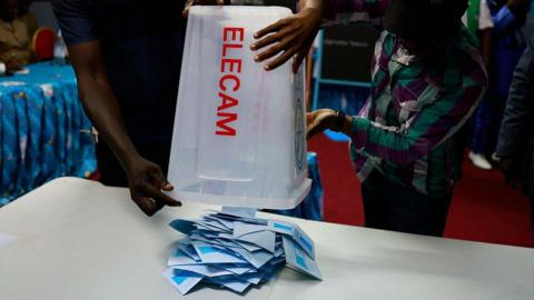 A pile of blue ballot slips are on a table below an upturned ballot box with the word "Elecam" written on it. Two people, who can't be seen in full, are holding the box upside down.