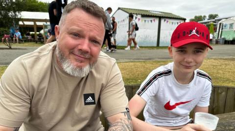 A man with short brown hair and a grey beard, wearing a beige Adidas t-shirt, sits at a picnic table next to a young boy wearing a white Nike t-shirt and a red baseball cap. The boy is holding a drink and both are smiling at the camera.