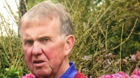 Head and shoulder shot of Malcolm Flynn, an elderly man with short grey hair. He is against a background of foliage with pink flowers at the bottom of the image.